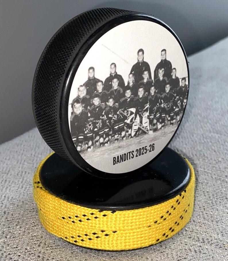 May include: A black hockey puck with a team photo and the text "BANDITS 2025-26" on a black base with yellow and black trim. The puck is standing upright, showcasing the team photo.