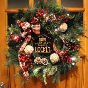 Op de afbeelding: Een kerstkrans met een geruit lint, rode bessen, dennenappels en een bord met de tekst "This home is filled with love, laughter, & hockey."