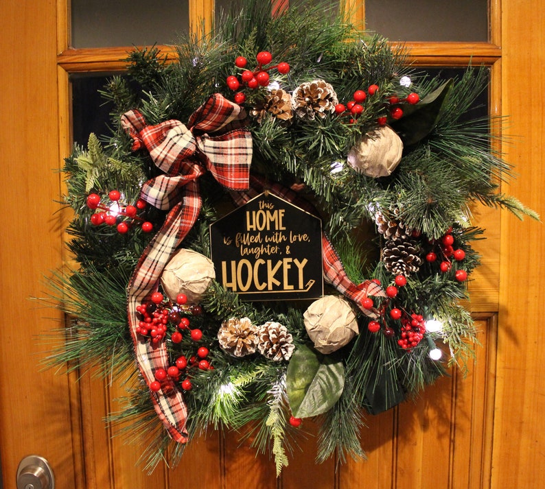 Op de afbeelding: Een kerstkrans met een geruit lint, rode bessen, dennenappels en een bord met de tekst "This home is filled with love, laughter, & hockey."