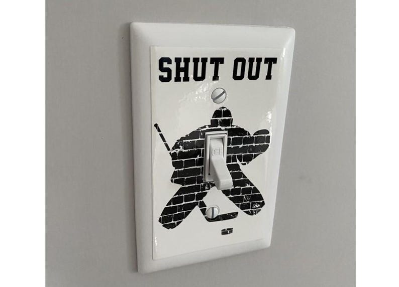 K&ouml;nnte beinhalten: Ein wei&szlig;er Lichtschalter mit einem schwarz-wei&szlig;en Aufkleber eines Hockeytorwarts vor einer Backsteinmauer. Der Aufkleber lautet "SHUT OUT".
