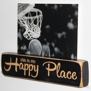 May include: Black wooden sign with the text "This is my Happy Place" in a light wood font. A black and white photo of a basketball hoop with a basketball in the net is displayed in a frame on top of the sign.