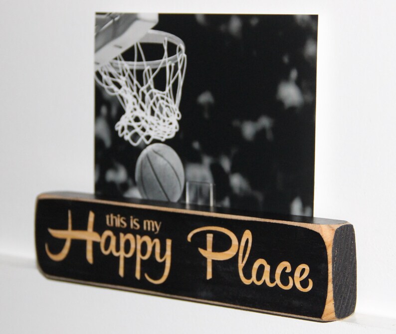 May include: Black wooden sign with the text "This is my Happy Place" in a light wood font. A black and white photo of a basketball hoop with a basketball in the net is displayed in a frame on top of the sign.
