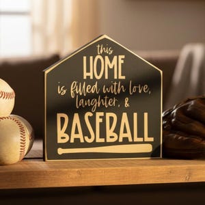 May include: A black, home plate-shaped wooden sign with gold lettering that reads: this HOME is filled with love, laughter, & BASEBALL, featuring a graphic of a baseball bat at the bottom, positioned next to two baseballs and a brown leather glove.