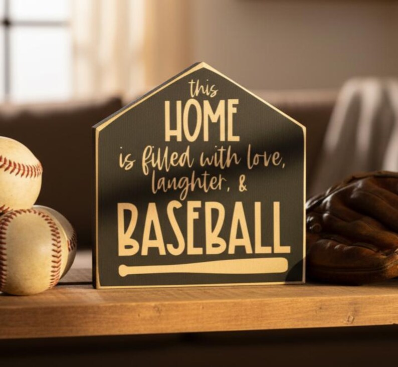 May include: A black, home plate-shaped wooden sign with gold lettering that reads: this HOME is filled with love, laughter, & BASEBALL, featuring a graphic of a baseball bat at the bottom, positioned next to two baseballs and a brown leather glove.