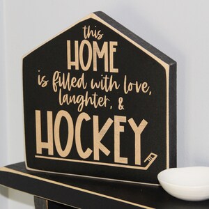 Op de afbeelding: Zwart houten bord in de vorm van een huis met de tekst "This home is filled with love, laughter, & hockey" in beige letters.