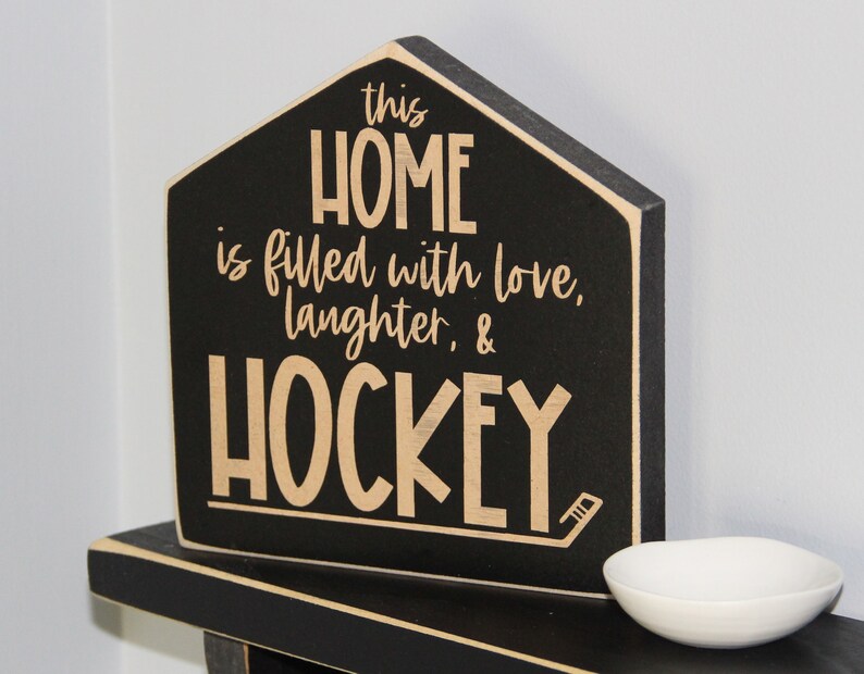 Op de afbeelding: Zwart houten bord in de vorm van een huis met de tekst "This home is filled with love, laughter, & hockey" in beige letters.