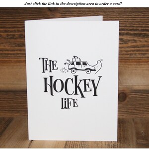 May include: A white card with black text and a simple illustration of a car with hockey sticks on top. The text reads "THE HOCKEY LIFE".