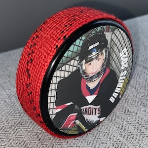 May include: A red hockey puck with a black border and a photo of a hockey player. The player is wearing a black and red jersey with the word "BANDITS" and the year "2025" printed on the puck.