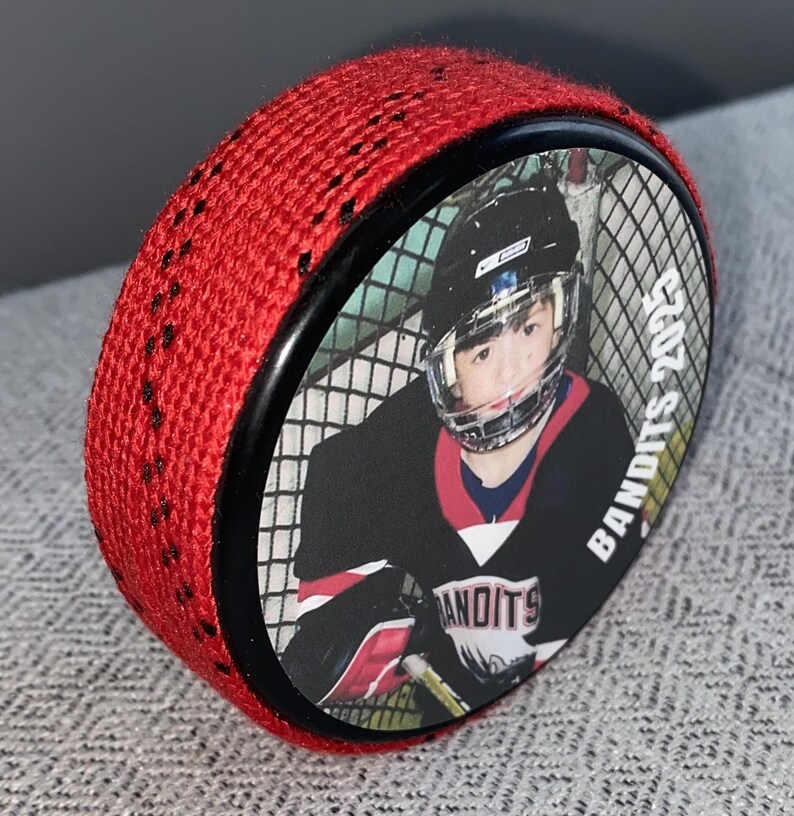 May include: A red hockey puck with a black border and a photo of a hockey player. The player is wearing a black and red jersey with the word "BANDITS" and the year "2025" printed on the puck.