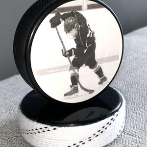 May include: A black hockey puck with a black and white image of a hockey player. The puck is mounted on a second puck, with white laces wrapped around the base. A unique sports-themed decorative item.