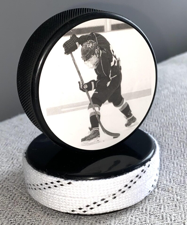 May include: A black hockey puck with a black and white image of a hockey player. The puck is mounted on a second puck, with white laces wrapped around the base. A unique sports-themed decorative item.