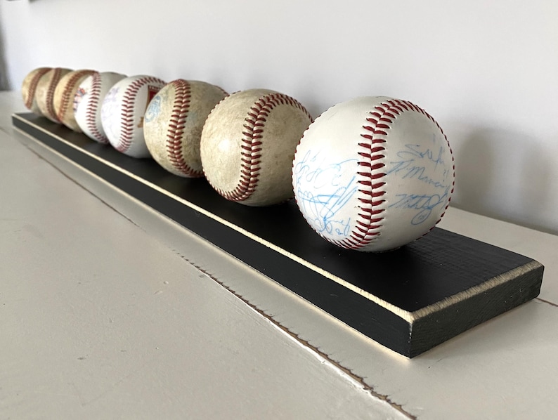 May include: A black wooden shelf displays six baseballs. The baseballs are white with red stitching and some have signatures. The shelf has a black top with a thin white border.