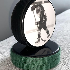 May include: A black hockey puck with a black and white image of a hockey player. The puck is mounted on a second puck with a green fabric wrap around the base. The image is of a hockey player in action.