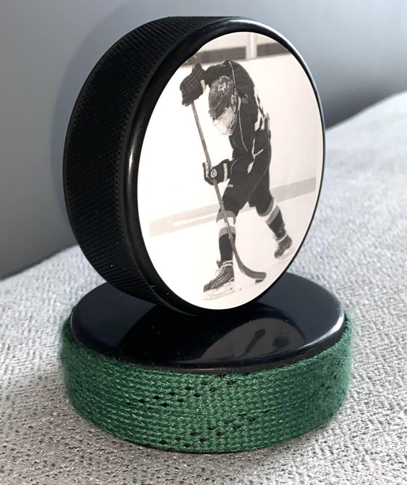 May include: A black hockey puck with a black and white image of a hockey player. The puck is mounted on a second puck with a green fabric wrap around the base. The image is of a hockey player in action.