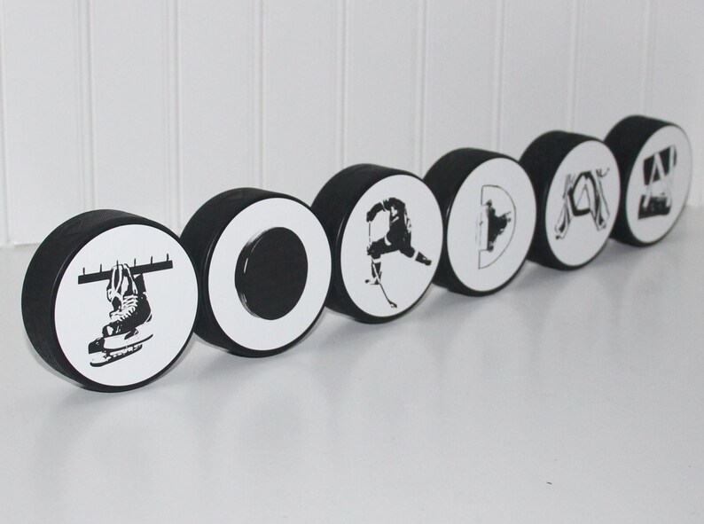 May include: A set of six black and white hockey puck ornaments spelling out the word "TORONTO". Each puck features a different hockey-themed design, including a hockey stick, a hockey player, and a hockey skate.