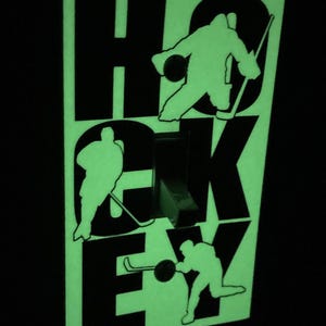 K&ouml;nnte beinhalten: Ein leuchtender Lichtschalter-Abdeckung, der im Dunkeln leuchtet, mit dem Wort "HOCKEY" in schwarzen Buchstaben. Jeder Buchstabe hat die Silhouette eines Hockeyspielers in einer Schlittschuh-Position.