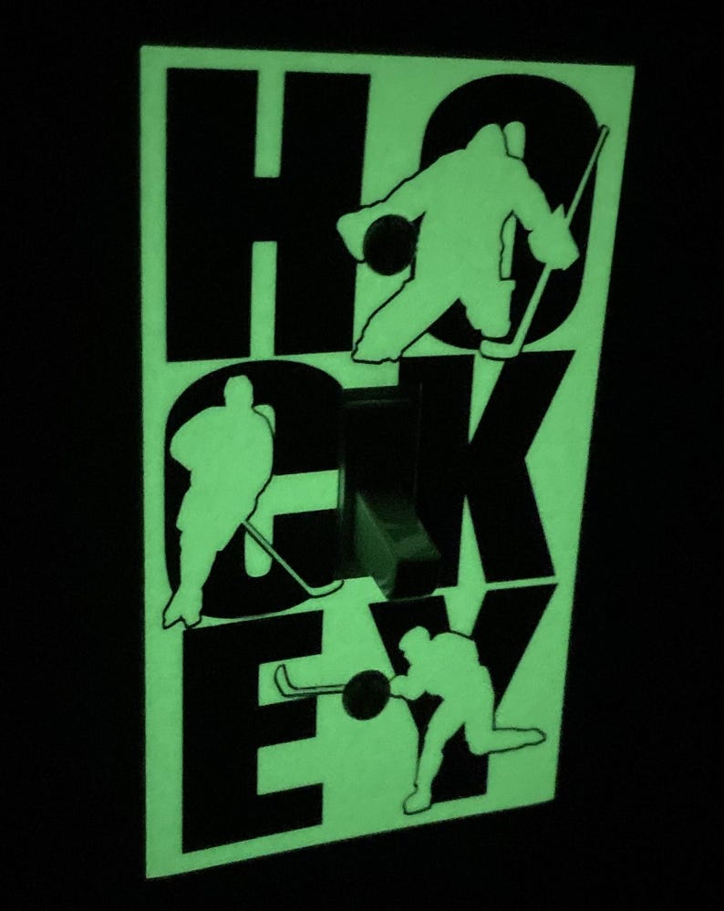K&ouml;nnte beinhalten: Ein leuchtender Lichtschalter-Abdeckung, der im Dunkeln leuchtet, mit dem Wort "HOCKEY" in schwarzen Buchstaben. Jeder Buchstabe hat die Silhouette eines Hockeyspielers in einer Schlittschuh-Position.