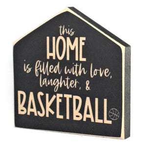 Op de afbeelding: Zwart houten bord in de vorm van een huis met de tekst "This home is filled with love, laughter, & basketball" in cr&egrave;mekleurige letters. Een klein basketbalicoon bevindt zich in de rechterbenedenhoek.