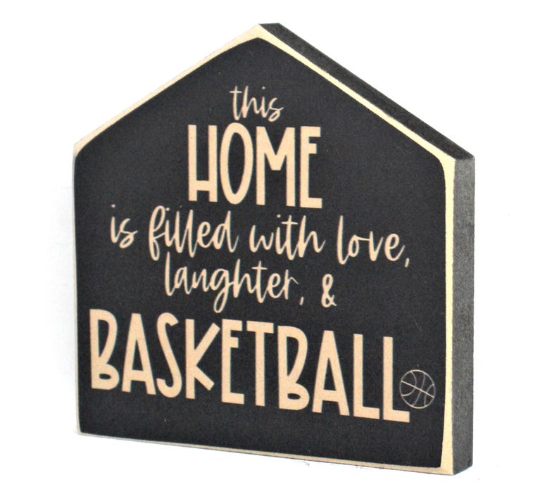 Op de afbeelding: Zwart houten bord in de vorm van een huis met de tekst "This home is filled with love, laughter, & basketball" in cr&egrave;mekleurige letters. Een klein basketbalicoon bevindt zich in de rechterbenedenhoek.