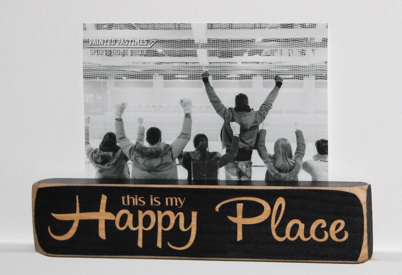 May include: Black wooden picture frame with the text "this is my Happy Place" in gold lettering. The frame holds a black and white photo of people cheering at a sporting event.