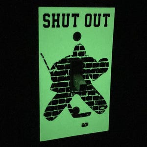 K&ouml;nnte beinhalten: Ein gr&uuml;ner Lichtschalter-Abdeckung mit einer schwarzen Silhouette eines Hockey-Torwarts vor einer Backsteinmauer. Der Text "SHUT OUT" ist oben auf der Abdeckung aufgedruckt.