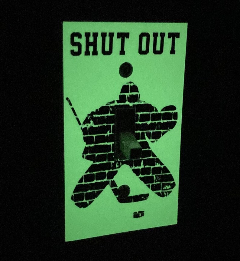 K&ouml;nnte beinhalten: Ein gr&uuml;ner Lichtschalter-Abdeckung mit einer schwarzen Silhouette eines Hockey-Torwarts vor einer Backsteinmauer. Der Text "SHUT OUT" ist oben auf der Abdeckung aufgedruckt.