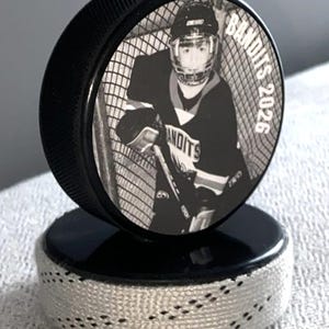 May include: A black hockey puck with a black and white image of a hockey player in a net. The text "Bandits 2026" is printed on the puck. The puck is on a black base with white stitching.