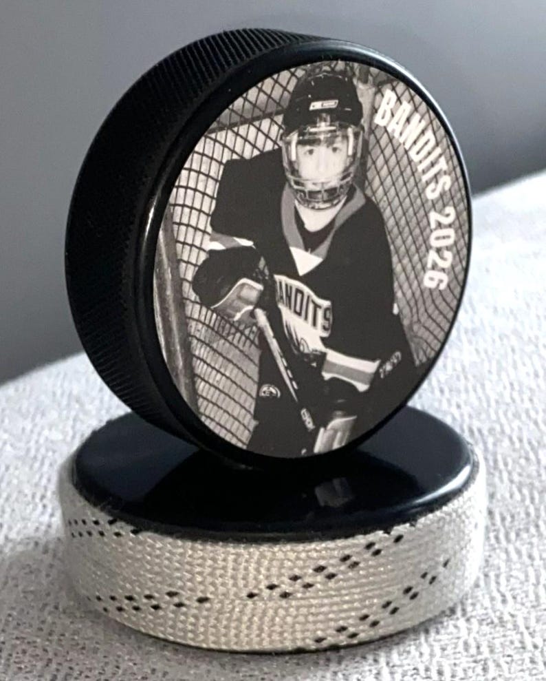 May include: A black hockey puck with a black and white image of a hockey player in a net. The text "Bandits 2026" is printed on the puck. The puck is on a black base with white stitching.