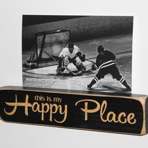 May include: Black wooden sign with the text "this is my happy place" in gold lettering. A black and white photo of hockey players on the ice is displayed above the sign.