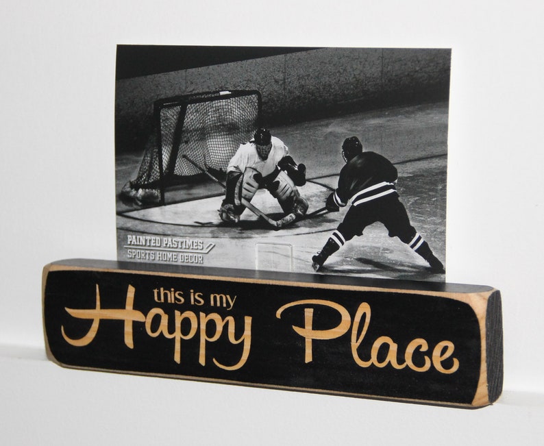 May include: Black wooden sign with the text "this is my happy place" in gold lettering. A black and white photo of hockey players on the ice is displayed above the sign.