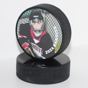 May include: A black hockey puck with a printed image of a hockey player in a black and red jersey. The puck has the text "2024 Bandits" and is displayed on top of another black puck.