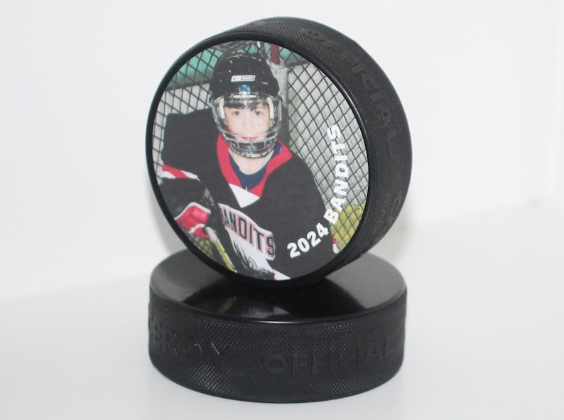 May include: A black hockey puck with a printed image of a hockey player in a black and red jersey. The puck has the text "2024 Bandits" and is displayed on top of another black puck.