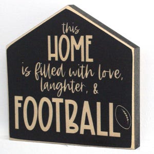 Op de afbeelding: Zwart houten bord in de vorm van een huis met de tekst "this HOME is filled with love, laughter, & FOOTBALL" in beige letters met een beige voetbalafbeelding.