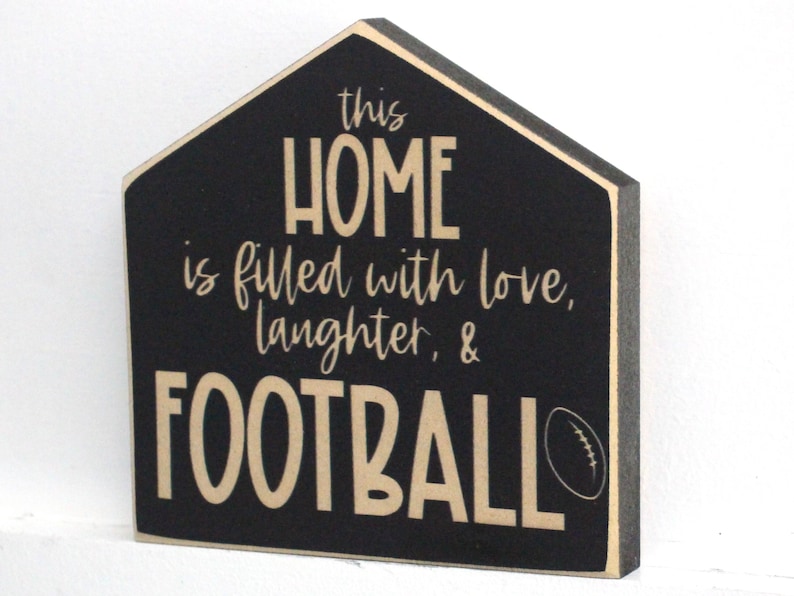 Op de afbeelding: Zwart houten bord in de vorm van een huis met de tekst "this HOME is filled with love, laughter, & FOOTBALL" in beige letters met een beige voetbalafbeelding.