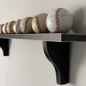 May include: A collection of baseballs with red stitching, displayed on a black wooden shelf. The shelf is mounted on a white wall. Some baseballs have signatures. The shelf is supported by two black brackets.