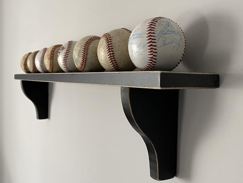 May include: A collection of baseballs with red stitching, displayed on a black wooden shelf. The shelf is mounted on a white wall. Some baseballs have signatures. The shelf is supported by two black brackets.