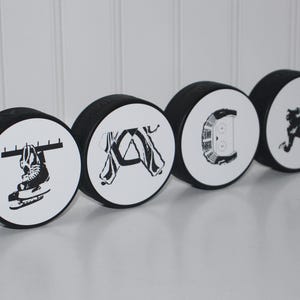 May include: Five black and white hockey puck ornaments with different hockey-themed designs. The designs include a hockey stick and puck, a hockey player, a hockey skate, a hockey net, and a hockey jersey.