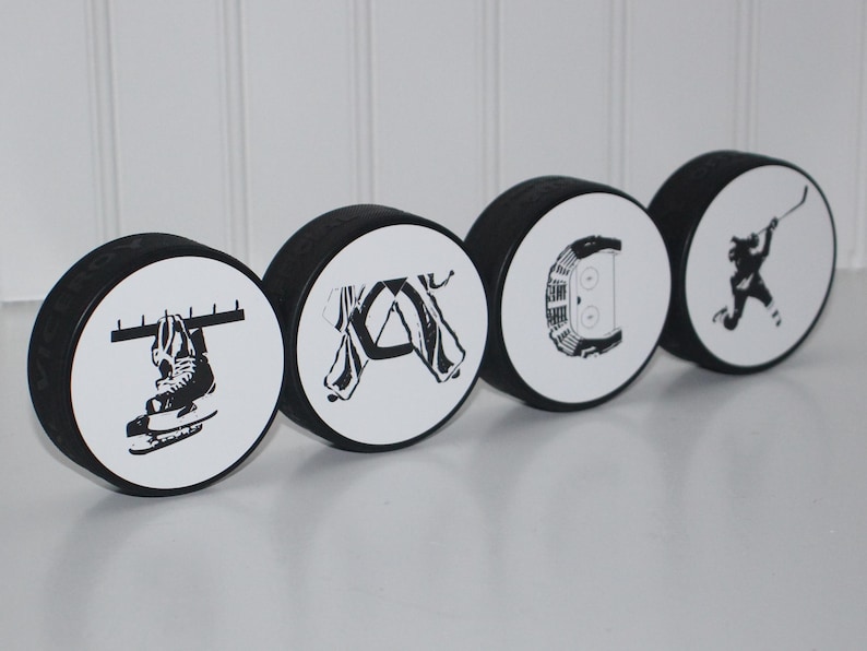 May include: Five black and white hockey puck ornaments with different hockey-themed designs. The designs include a hockey stick and puck, a hockey player, a hockey skate, a hockey net, and a hockey jersey.
