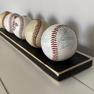 May include: A display of five baseballs with red stitching and signatures, resting on a black wooden base with a white trim. The baseballs are arranged in a row, showcasing their details.