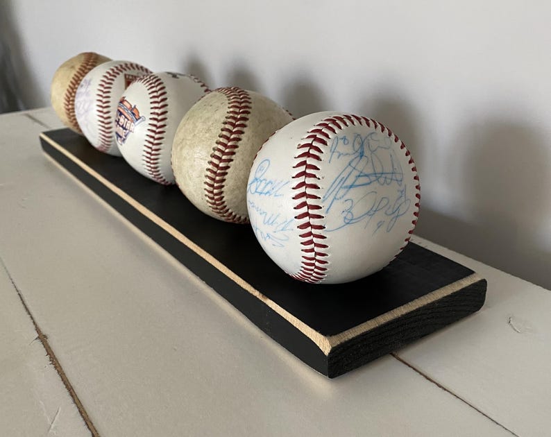 May include: A display of five baseballs with red stitching and signatures, resting on a black wooden base with a white trim. The baseballs are arranged in a row, showcasing their details.