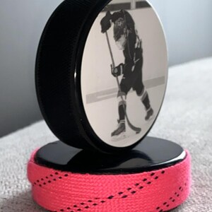 May include: A black hockey puck with a laser-etched image of a hockey player, mounted on a black base with a bright pink shoelace wrap. The puck is angled, showcasing the player in action. A unique sports-themed decorative item.