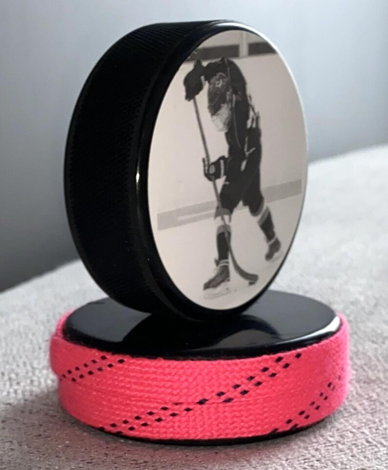 May include: A black hockey puck with a laser-etched image of a hockey player, mounted on a black base with a bright pink shoelace wrap. The puck is angled, showcasing the player in action. A unique sports-themed decorative item.