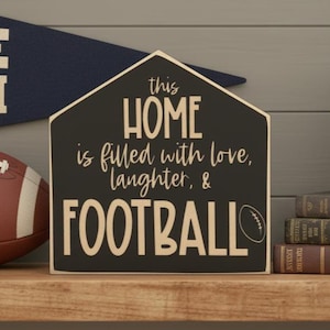 May include: A house-shaped wooden sign with a black background and cream-colored text that reads: this HOME is filled with love, laughter, & FOOTBALL. The sign features a small graphic of a football next to the word football in a bold, sans-serif font.