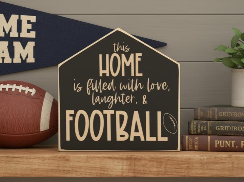 May include: A house-shaped wooden sign with a black background and cream-colored text that reads: this HOME is filled with love, laughter, & FOOTBALL. The sign features a small graphic of a football next to the word football in a bold, sans-serif font.