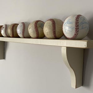 May include: A collection of baseballs with red stitching and varying degrees of wear, displayed on a cream-colored wooden shelf. The shelf is supported by two decorative brackets, creating a rustic display.