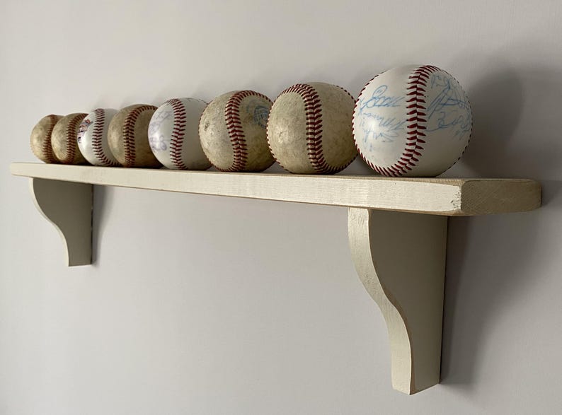 May include: A collection of baseballs with red stitching and varying degrees of wear, displayed on a cream-colored wooden shelf. The shelf is supported by two decorative brackets, creating a rustic display.