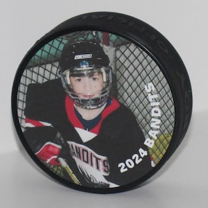 May include: Black hockey puck featuring a color image of a hockey player in a helmet and jersey. The jersey has the word "Bandits" and the year "2024" printed on it. The puck is a standard size for ice hockey.