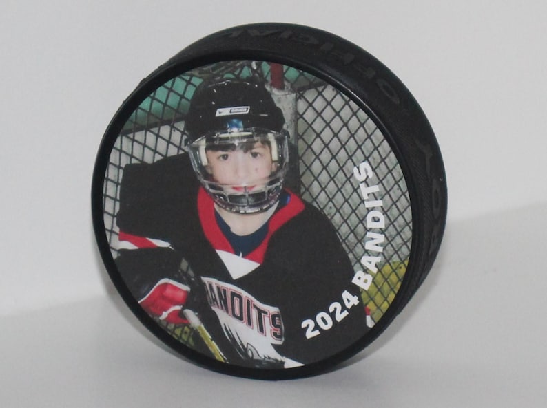 May include: Black hockey puck featuring a color image of a hockey player in a helmet and jersey. The jersey has the word "Bandits" and the year "2024" printed on it. The puck is a standard size for ice hockey.