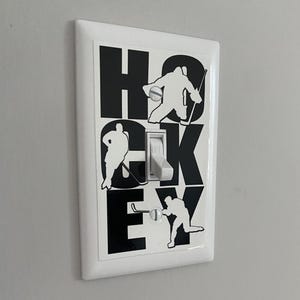 K&ouml;nnte beinhalten: Ein wei&szlig;er Lichtschalter mit einem schwarz-wei&szlig;en Aufkleber von Hockeyspielern und dem Wort "HOCKEY".