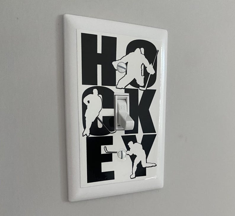 K&ouml;nnte beinhalten: Ein wei&szlig;er Lichtschalter mit einem schwarz-wei&szlig;en Aufkleber von Hockeyspielern und dem Wort "HOCKEY".